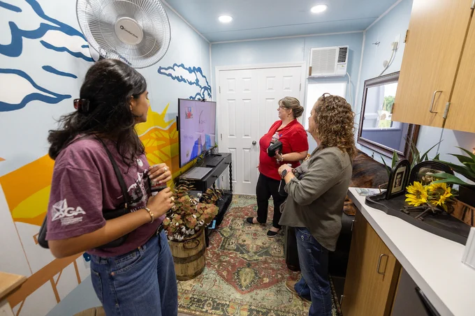 A group of people stand in a small, colorful room with painted walls and an indoor plant. They are focused on a television displaying a videoconference with a health professional. The room features a patterned rug, wooden cabinets and a large fan on the ceiling.