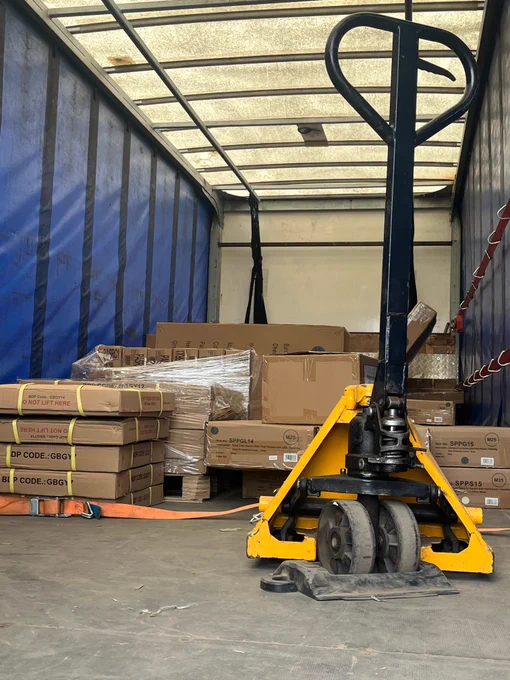 Yellow pallet jack positioned inside a truck trailer loaded with stacked cardboard boxes and wrapped pallets.
