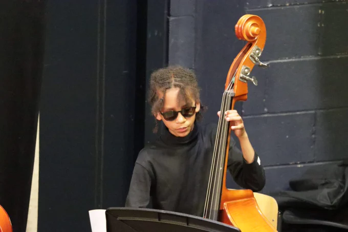 Young musician wearing sunglasses playing a double bass on stage against a dark brick wall background.