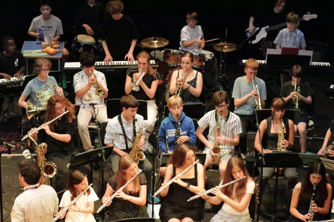 A diverse group of young musicians performing on stage with various instruments including flutes, saxophones, trumpets, and keyboards.