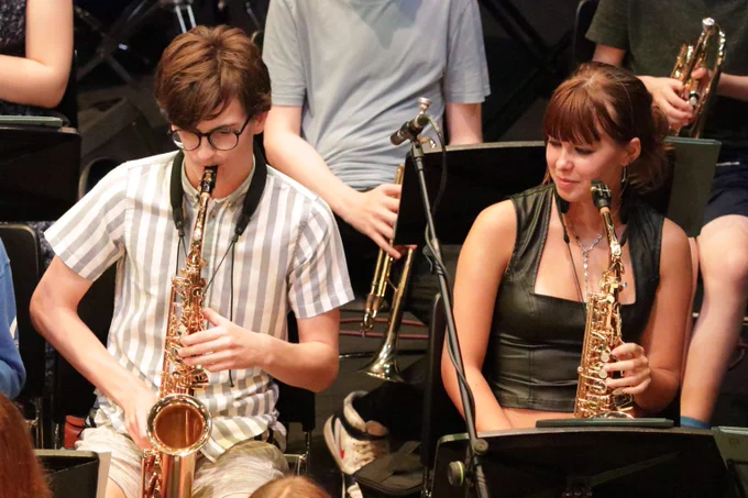 Two young musicians playing saxophones seated in a band setting with music stands in front of them.