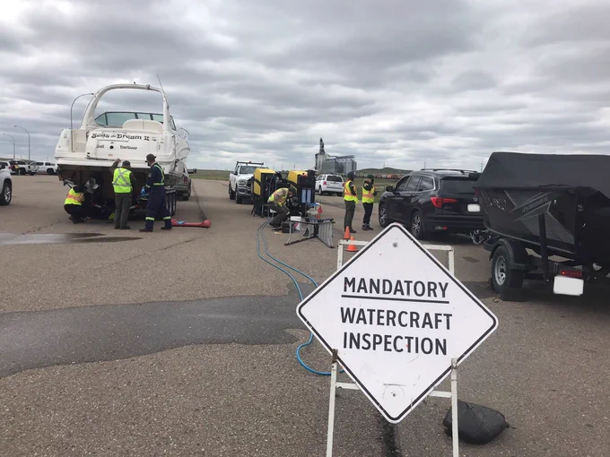 A sign in the foreground says Mandatory Watercraft Inspection. In the background, trailered boats are stopped at the station while inspectors search watercraft for signs of invasive species. The sky is cloudy and the pavement is wet.