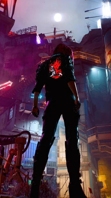 V poses with her back to the viewer in front of a tall, industrial building. It is made up of stacked storage containers joined by railings and stairs. V wears a jacket with a glowing Samurai logo on the back of an oni mask with cybernetic implants on half of its face.