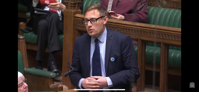A man in a suit and tie speaking in a parliamentary chamber with green leather benches and wooden details.