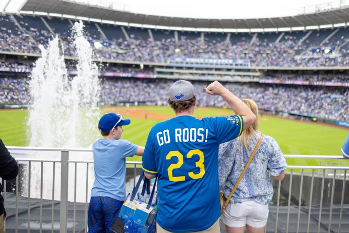 A family stands at the fence behind fountains at Kauffman Stadium, looking out at the baseball field. One person is wearing a UMKC/Royals jersey.