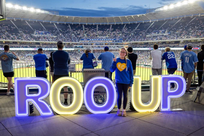 Light up letters spell out "ROO UP" in blue and gold on the concourse at Kauffman Stadium. Fans stand at the fence behind the letters, watching the game from behind the outfield.