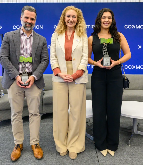 Three people stand smiling, each holding an award. The awards say \"text@1 Mobility Champion text@2\". The backdrop is blue with text that says \"CO text@3 MO text@4\".