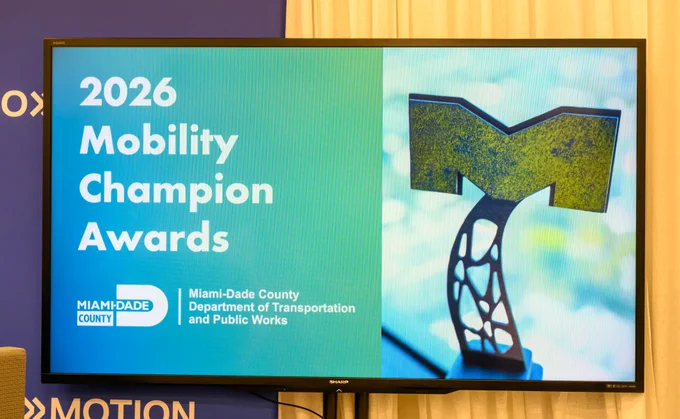 Display showing \"2026 Mobility Champion Awards\". Display also shows Miami-Dade County Department of Transportation and Public Works logo and an abstract 'M' statue. The display brand is SHARP AQUOS. \u00BBMOTION shown at the bottom.