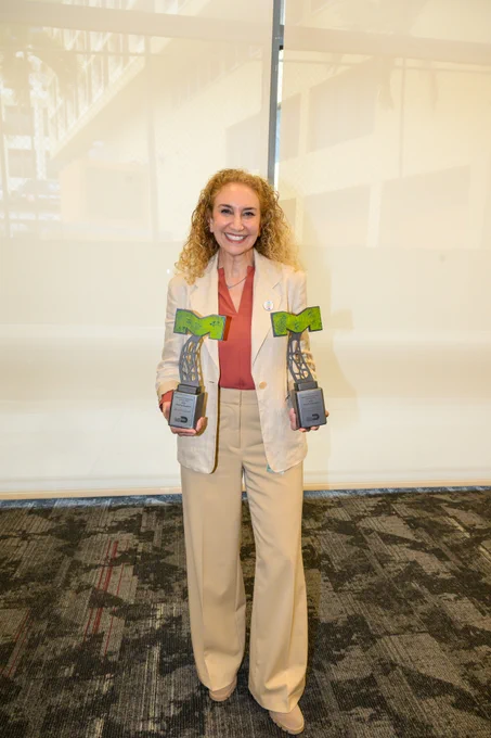 A woman with curly hair smiles, holding two awards. The awards read, \"Innovative Transportation and Public Works 2024 Mobility Champion, The FRED Group.\"
