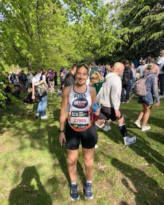 London Marathon runner wearing a race vest and bib, standing in a grassy park area holding a water bottle, with other participants around.