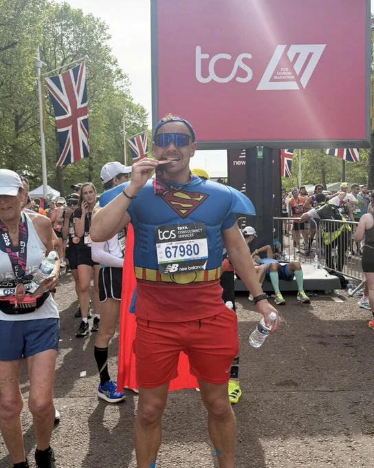 London Marathon finisher wearing a Superman outfit and medal, standing beneath a TCS London Marathon sign.