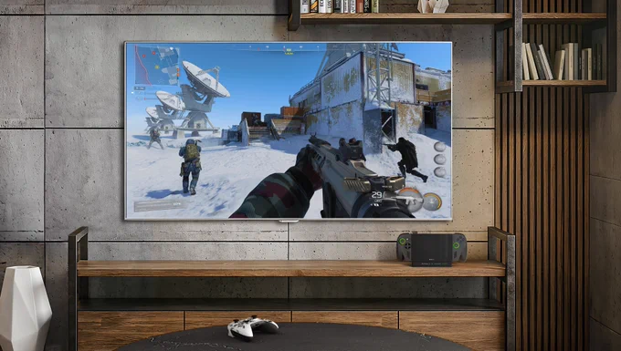 Static image of a TV set up with a video game shown on screen. The ROG Xbox Ally sits underneath the TV on a TV console. An Xbox controller lays on the coffee table. There are bookshelves above and to the right of the TV.