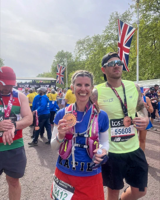 London Marathon runner standing near the finish area, holding a finisher medal and Union Jack flags in the background.”