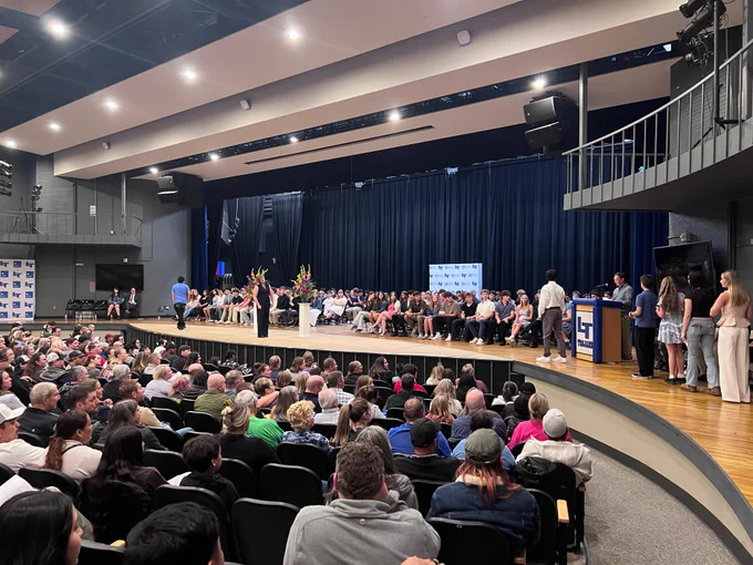 A large school auditorium filled with an audience seated in curved rows of chairs. On the raised stage, rows of students are seated while a presenter in a black outfit stands at a microphone. A podium with the "LT Trojans" logo is visible at right, where additional students and staff stand. Floral arrangements decorate the stage. Sponsor banners with the "LT" logo line the back wall.