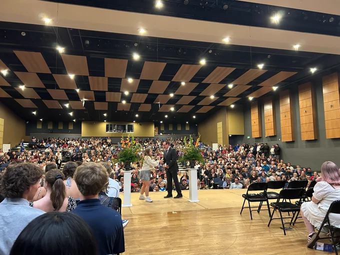 A packed school auditorium during an awards ceremony. A male presenter in a dark suit shakes hands with a female student at center stage, flanked by large floral arrangements on white pedestals. Hundreds of audience members fill the floor and bleachers in the background. Wooden acoustic panels line the walls and ceiling.