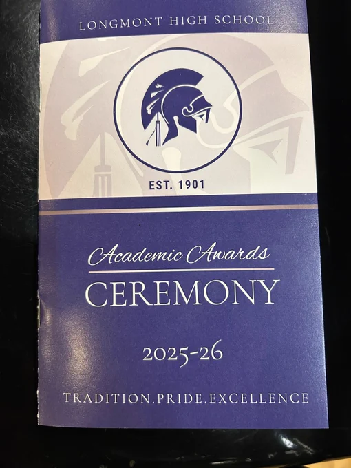 The cover of a printed program booklet for the Longmont High School Academic Awards Ceremony, 2025–26. The top half features a blue and white design with the school's Trojan helmet logo inside a circle and the text "Est. 1901." The bottom half is dark blue with white script and serif text reading "Academic Awards Ceremony 2025–26" and the school motto "Tradition. Pride. Excellence."