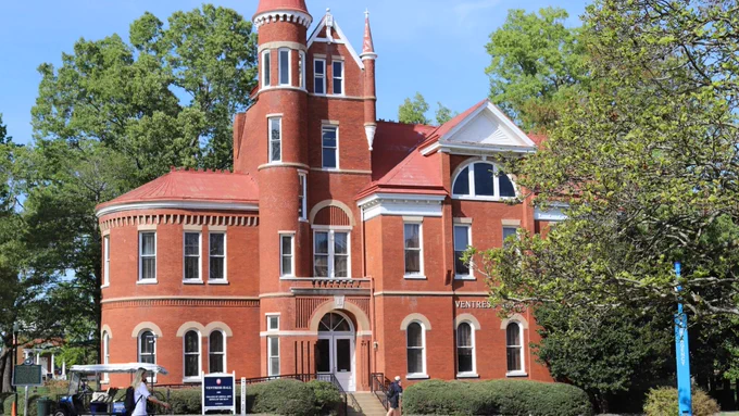 A red brick building with Victorian-style architecture, featuring a turret and arched windows, surrounded by trees. A small sign nearby reads "Ventress Hall."