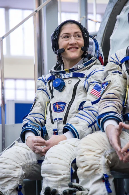 Female astronaut in a white space suit with American flag patch sitting indoors, hands resting on knees, preparing for a mission.
