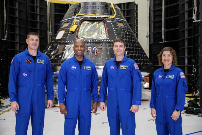 Four astronauts in blue NASA flight suits stand in front of a space capsule inside a large hangar.