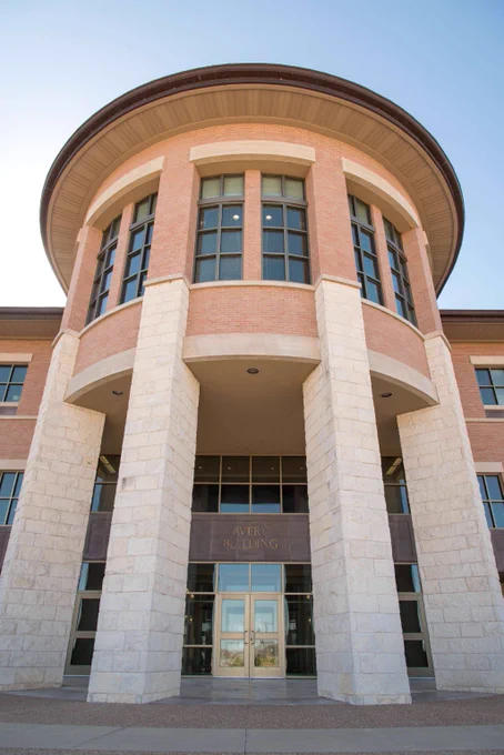 Tall round entrance of a modern building with large stone pillars and multiple windows under a clear blue sky.