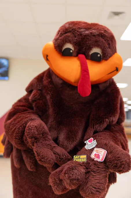 The HokieBird holding 3 new Cheesy Nights sticker designs. One sticker says, "Say cheese!" Another one says, "I love Cheesy Nights." The 3rd one is a lunch box design with the Newman Library building on the front. 