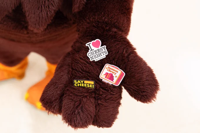The HokieBird's hand holding 3 new Cheesy Nights sticker designs. One sticker says, "Say cheese!" Another one says, "I love Cheesy Nights." The 3rd one is a lunch box design with the Newman Library building on the front. 