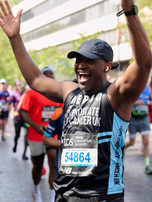 A person running in the London Marathon, wearing a Prostate Cancer UK running top. The person is smiling and has arms raised in a celebratory gesture.