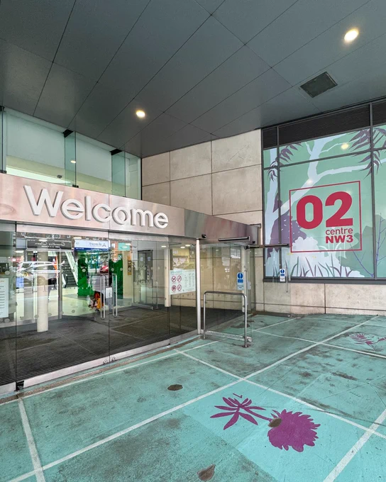 The image shows the entrance to the O2 Centre NW3, with a glass door beneath a "Welcome" sign. A large wall displays the O2 Centre NW3 logo with stylized green and purple foliage graphics.