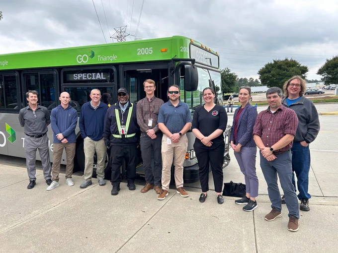 A group of people stands in front of a green and gray GO Triangle bus with "Special" displayed on the front sign. The group is smiling, and the bus is parked on a concrete surface with trees and a cloudy sky in the background.