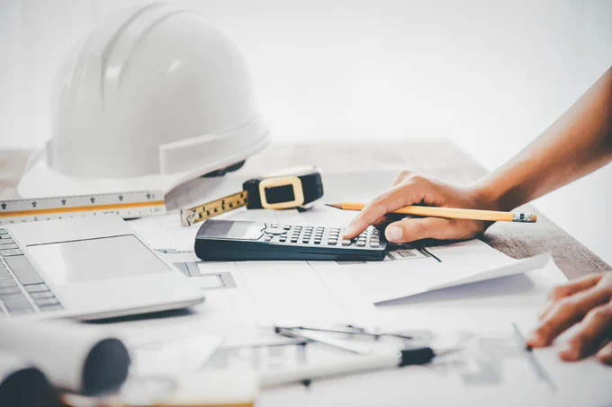 A person uses a calculator on a desk filled with architectural plans and tools, including a ruler, protractor, tape measure, and a white hard hat.