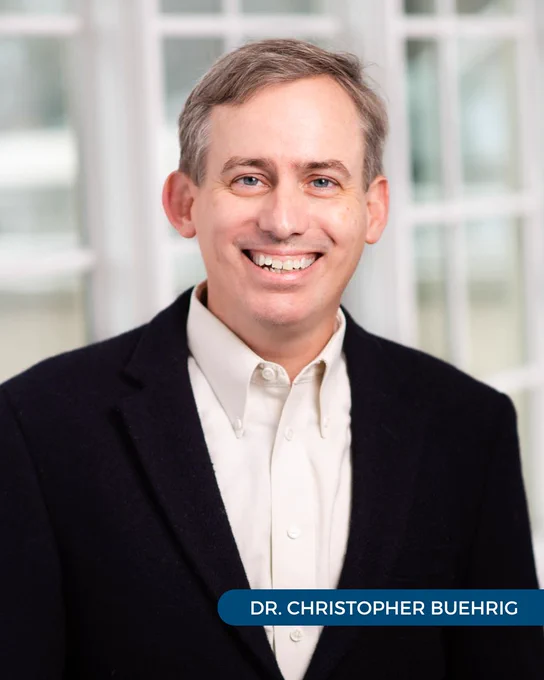 Dr. Christopher Buehrig of Tryon Medical Partners smiles and wears a dark blazer in a professional portrait in front of a paned glass window