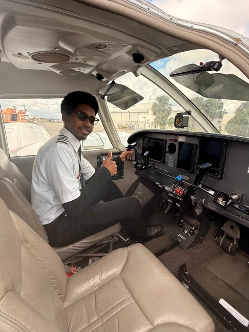 A person seated in the cockpit of a small aircraft is giving a thumbs-up gesture. The cockpit features various controls and instruments. In the background, other planes and a building can be seen. The person is wearing a uniform with epaulettes.