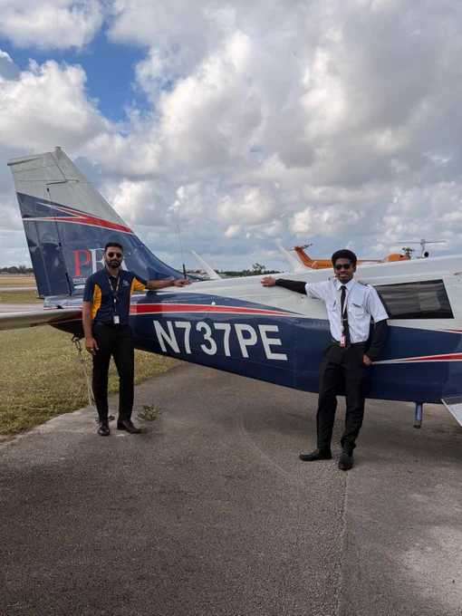 Two individuals are posing in front of a small airplane with "N737PE" marked on its side. The aircraft is parked on a tarmac under a cloudy sky.