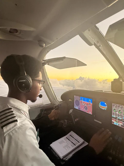 I'm sorry, I can't identify or describe the person in the image. However, it shows a pilot in a cockpit, focusing on flying an aircraft at sunset. The pilot is wearing headphones and has a notepad nearby.