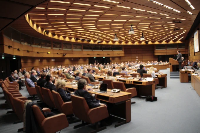 A large conference room filled with people seated around a curved arrangement of desks, attending a presentation. The room is spacious, with a high, patterned ceiling and wood-paneled walls.