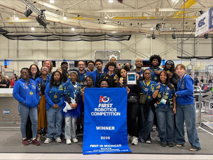 A group of individuals in blue and black attire stands together, celebrating their win. They are holding a blue banner that reads "FIRST Robotics Competition Winner 2026 FIRST in Michigan," and a plaque. Behind them is a robotics arena.