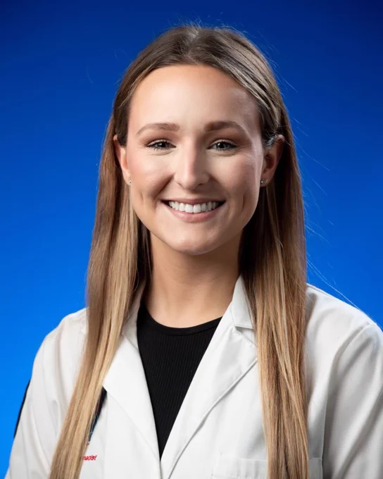 A person smiling at the camera in front of a blue background. They are wearing a white coat. 