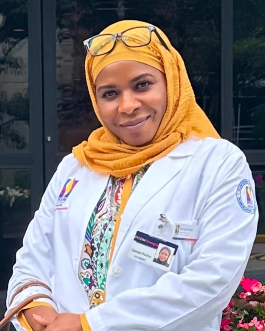 A person is wearing a white lab coat with a colorful patterned top underneath. The lab coat has a badge and a pair of glasses rests on the person's head. The background includes a building facade and some flowers.