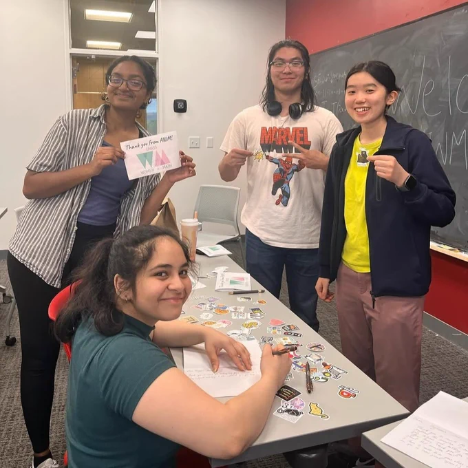 Four students standing and sitting around a table at an AWM Student Chapter event with a blackboard in the background. One student is seated at a table with colorful stickers arranged on the table. One of the standing students is holding up an AWM sign. All 4 students are smiling.