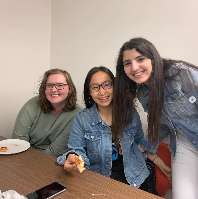 Three women seated at a classroom table eating pizza at an AWM Student Chapter event.