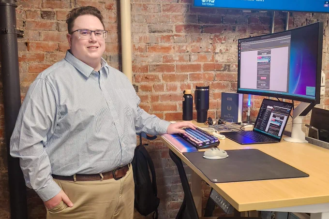 Man in a light blue shirt and khaki pants standing next to a desk with dual monitors and a laptop in a brick-walled office.