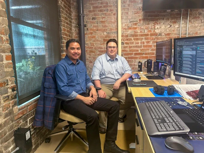 Two men sitting in an office with exposed brick walls, working at a desk with multiple computer monitors and keyboards.