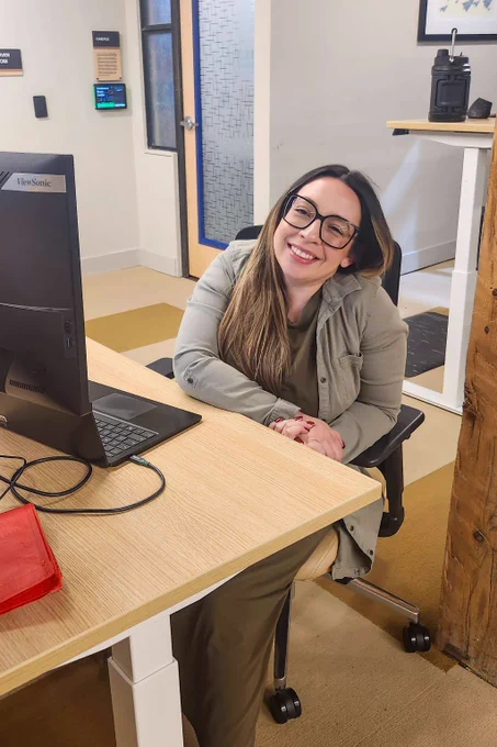 Woman seated at an office desk with a computer monitor and laptop, hands folded, in a modern workspace with light walls and wooden accents.