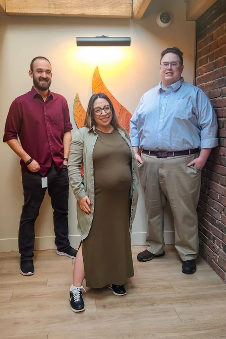 Three adults standing indoors against a wall with a decorative flame-shaped light fixture behind them.