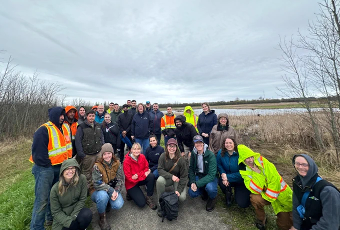 A group of people in outdoor clothing and reflective vests are gathered on a grassy area near a body of water with reeds. The sky is overcast. The setting appears to be a nature reserve or park.