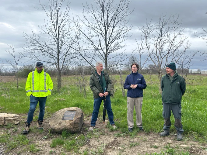 Four people stand outdoors near a large rock with a plaque, in front of leafless trees. One person wears a bright yellow jacket, and the others are in darker colors. The setting appears to be a natural area with overcast skies.