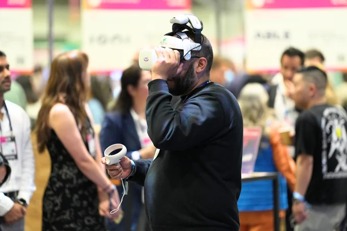A person wearing a virtual reality headset and holding a controller, with several people talking in the background.