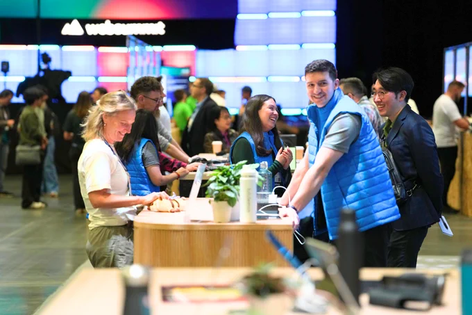 Busy event with attendees interacting and smiling at a wooden counter with plants and electronic devices, and a “Web Summit” logo in the background.