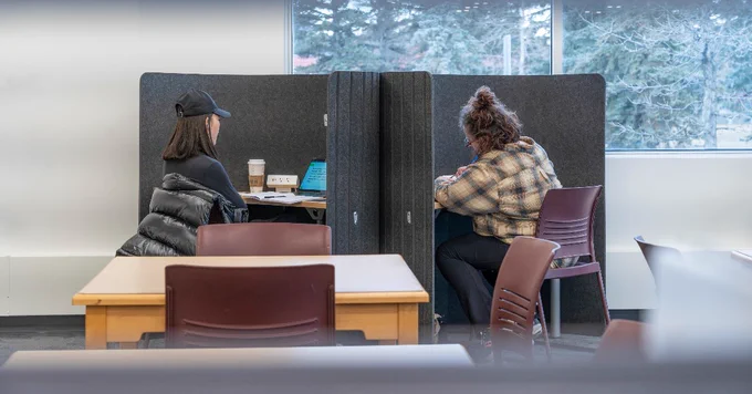 Two people are sitting in a study area with partitioned desks. One is wearing a cap and using a laptop with a coffee cup nearby, while the other is writing.