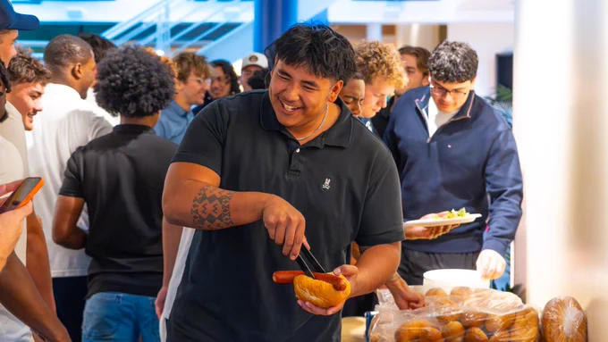 UC Davis student athlete has big smile on his face while he puts a sausage into a bun at a dinner on the food line